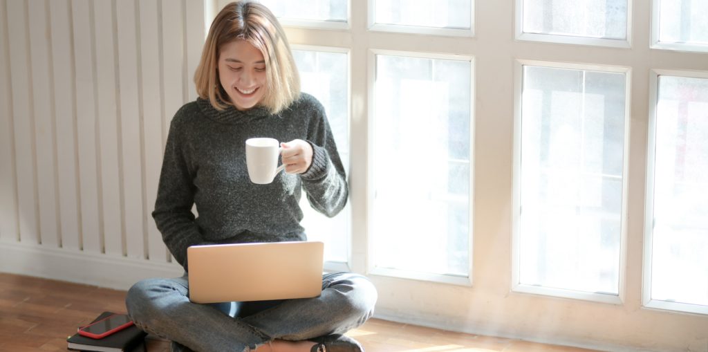 Picture of a young woman sitting on the floor, using her laptop, smiling with a cup of coffee